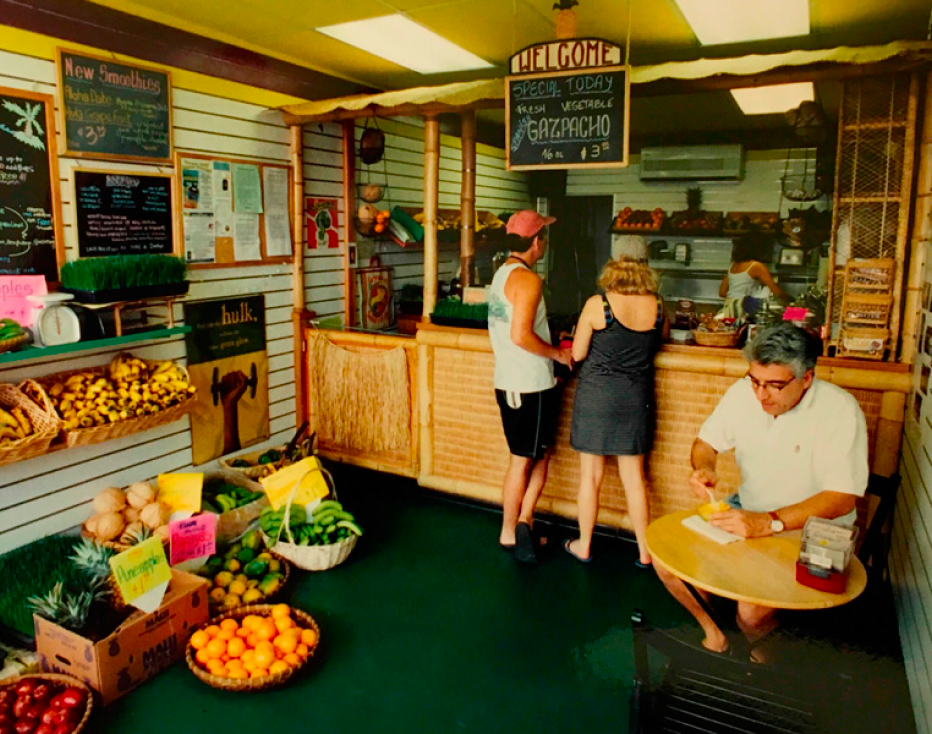 Photo in the interior of a Lanikai Juice with customers at the counter and basket of fruits on the floor.